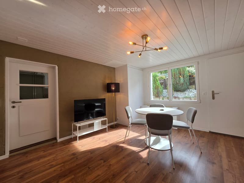 Bright, modern room with wooden flooring, a round dining table with four chairs, a television on a stand, a floor lamp, and a chandelier with multiple lights.
