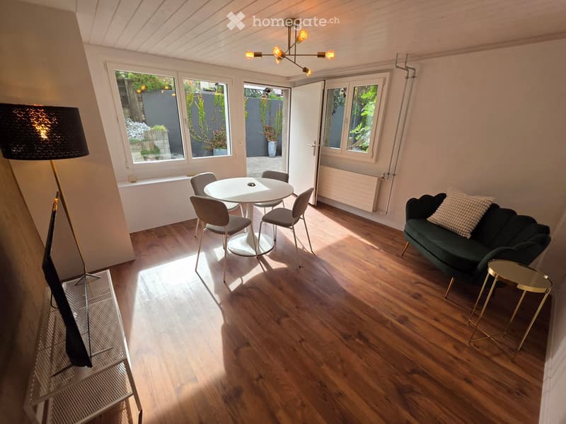 Bright living room with wooden flooring, large windows, a dark green sofa, a white round dining table with four chairs, a tall floor lamp, and a small gold side table.