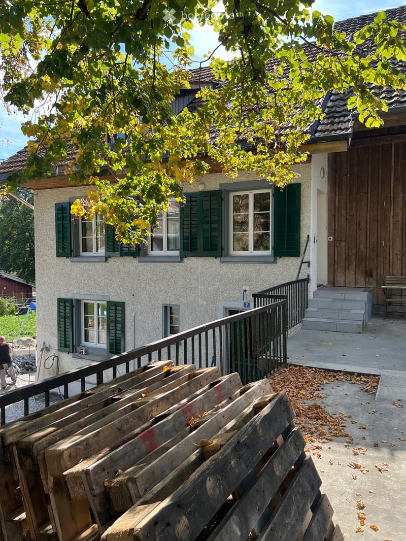 A two-story house with green shutters and a wooden balcony, surrounded by autumn leaves and stacked wooden planks on the ground.