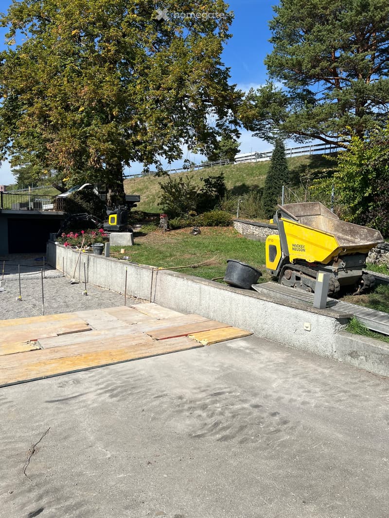 Outdoor construction area with concrete slabs, wooden planks, construction equipment, and a raised ground floor.