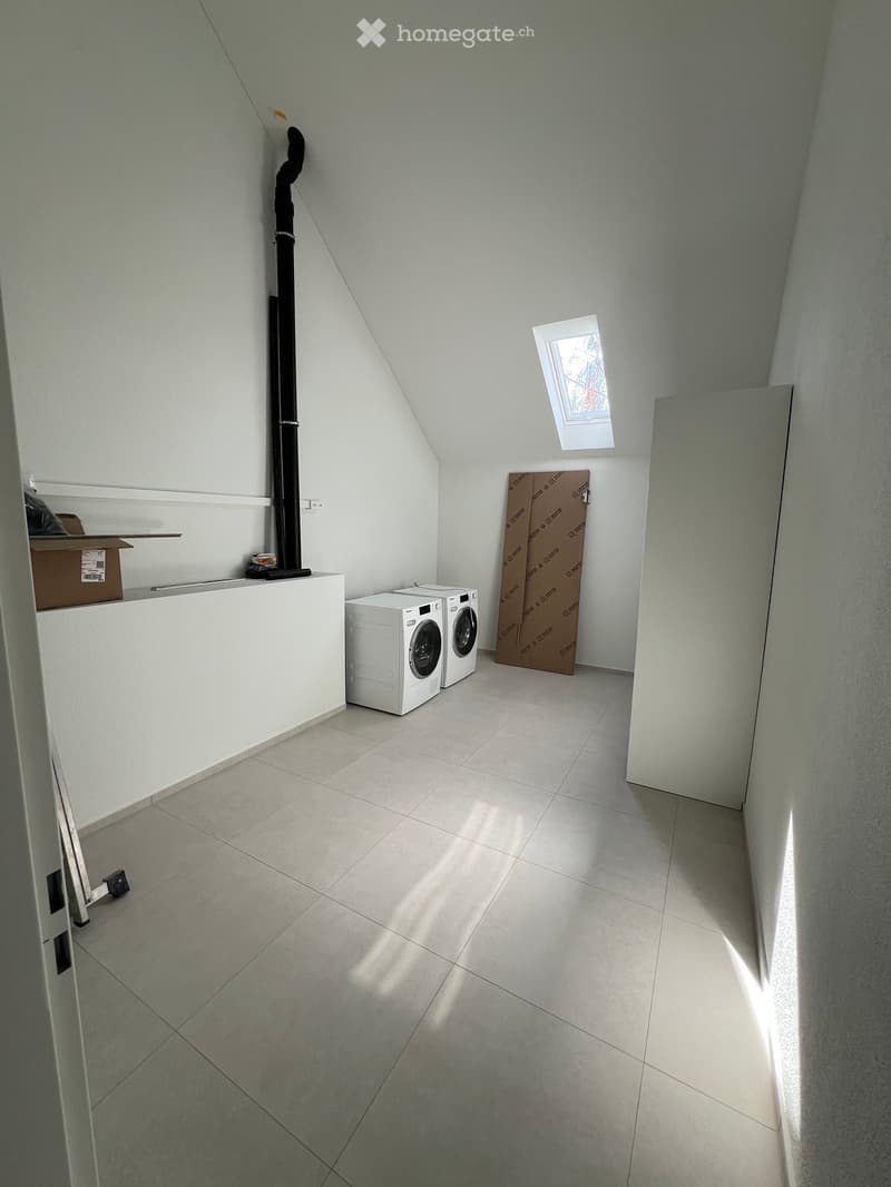 Bright, clean room with tiled floor, white walls, and a skylight window. Contains a washing machine and dryer, with a small shelf and cardboard boxes visible.