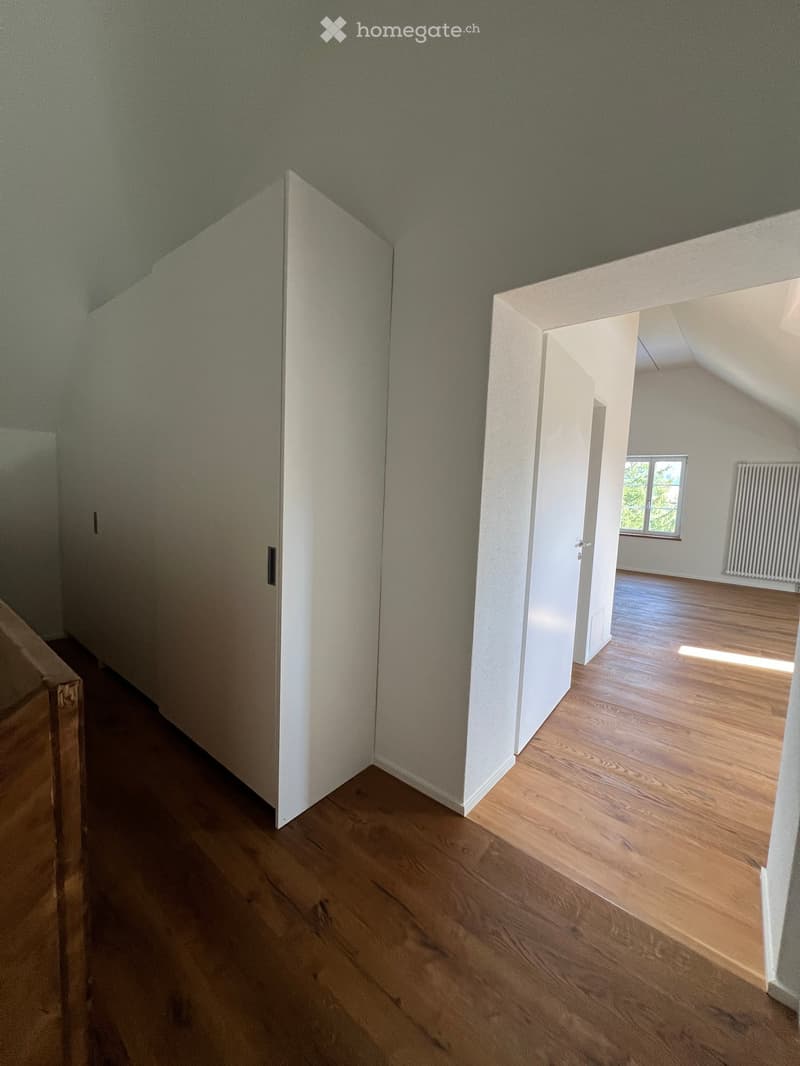 Empty hallway with wooden flooring, white walls, and a door leading to another room with a window and radiator.