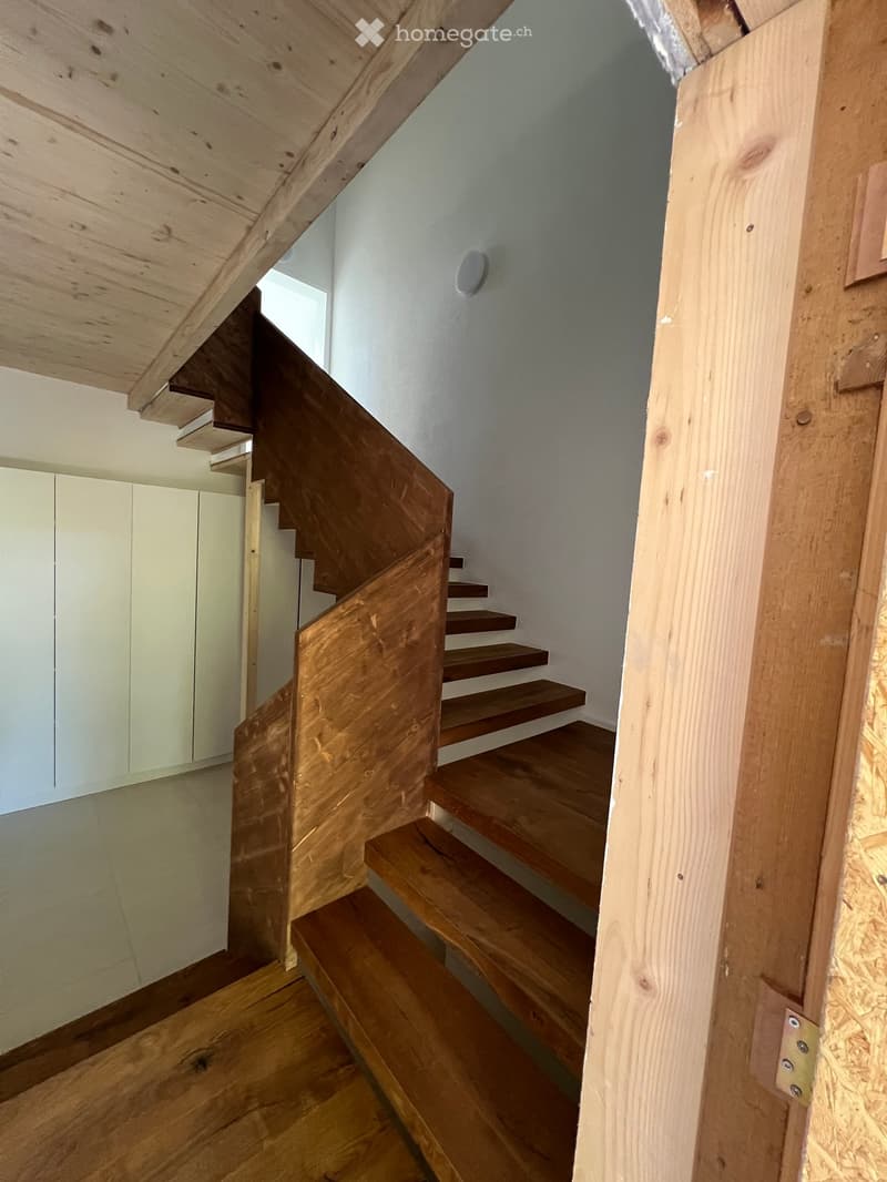 Wooden staircase with a modern design, featuring a sleek wooden railing and steps, leading upwards with a visible light fixture on the wall.