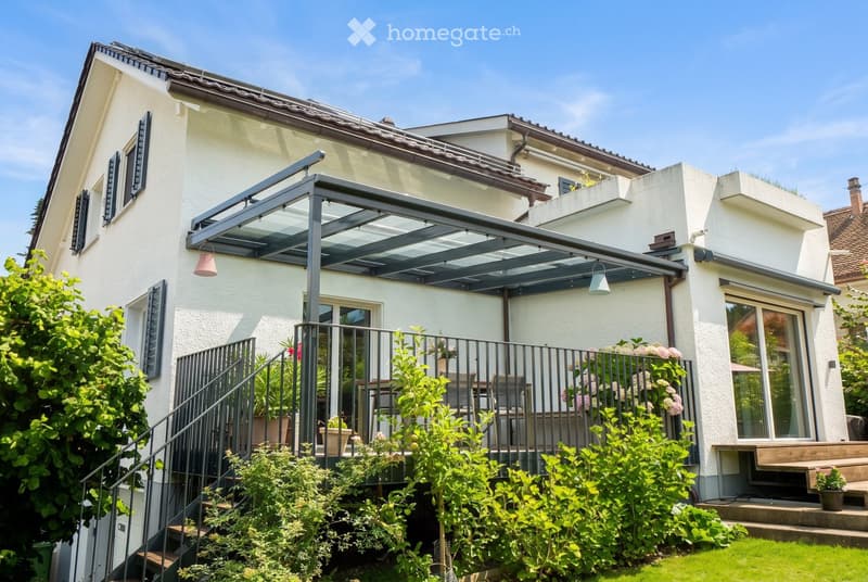A modern two-story house with a covered patio area, featuring a staircase leading up to a balcony, surrounded by greenery and potted plants.