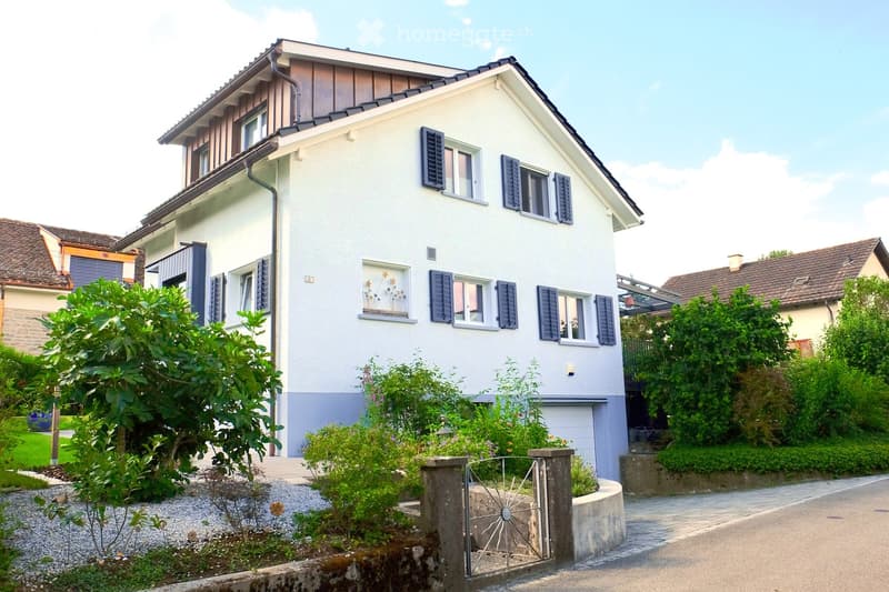 Two-story house with white exterior, dark shutters, and a garage. The house features a well-maintained garden with various plants and a decorative metal gate.