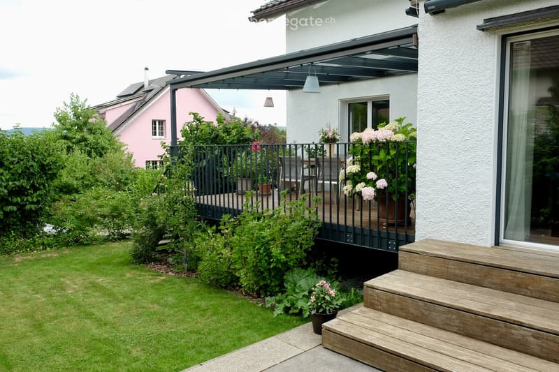 Well-maintained garden with a wooden staircase leading to a covered balcony area, surrounded by greenery and potted plants.