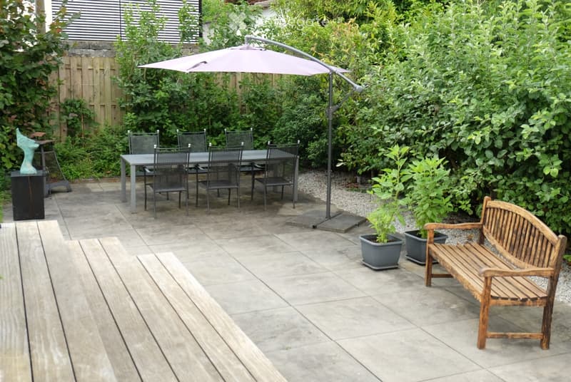 A well-maintained garden patio with a dining table, chairs, a wooden bench, potted plants, and a large umbrella providing shade. The area is enclosed by greenery and features a stone pathway and wooden decking.