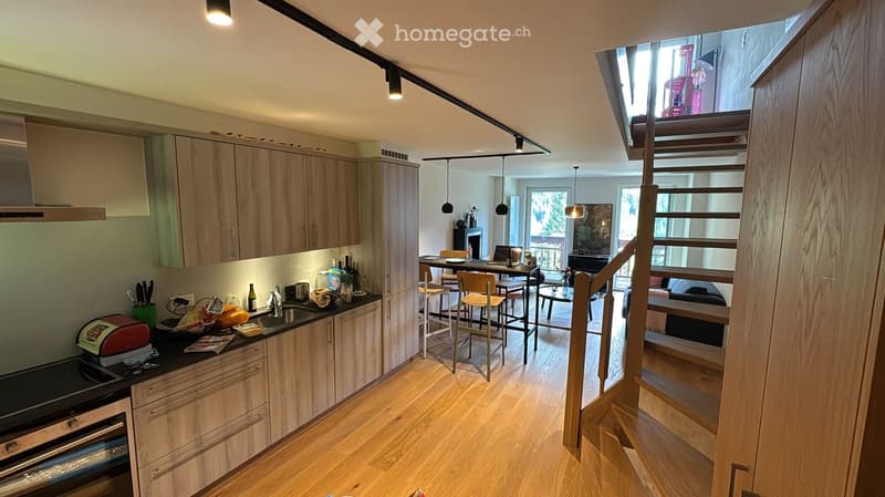 Open-plan kitchen and living area with light wood cabinets, stainless steel appliances, a dining area with stools, and a living area with a sofa and piano.