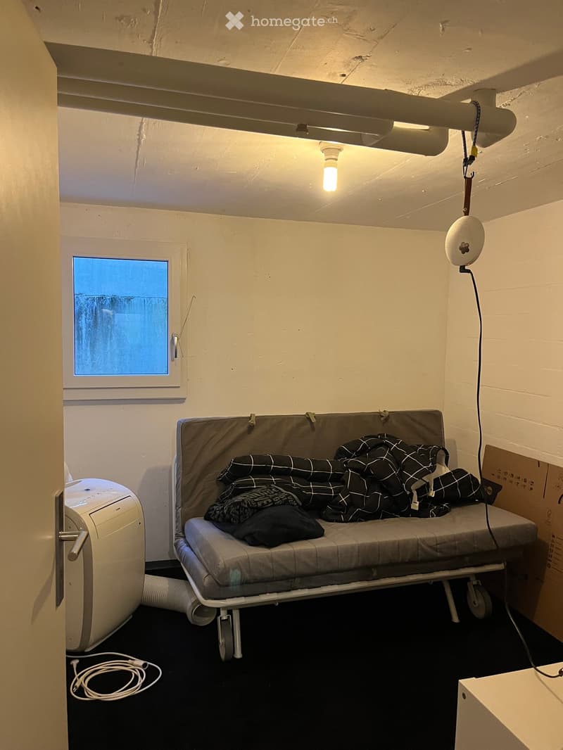 Small room with a sofa, blanket, air conditioner, and a speaker. The room has a window and a light fixture on the ceiling.