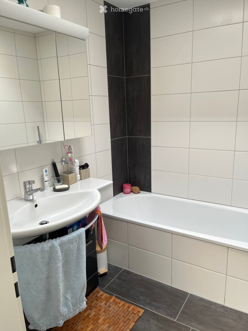 Modern bathroom with white tiles, a white sink with a chrome faucet, a bathtub, and a towel hanging on a rack. The floor has dark tiles with a wooden bath mat.