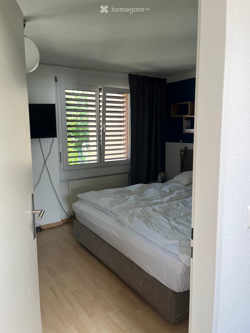 Bedroom with a double bed, white bedding, wooden floor, radiator, shelves, and a window with blinds.