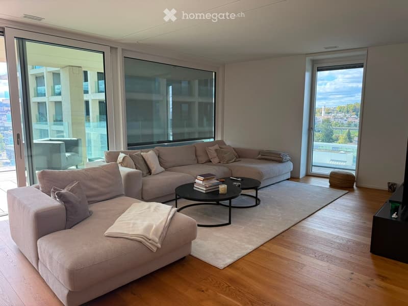 Modern living room with large windows, light-colored sectional sofa, coffee table, ottoman, and books on the table.
