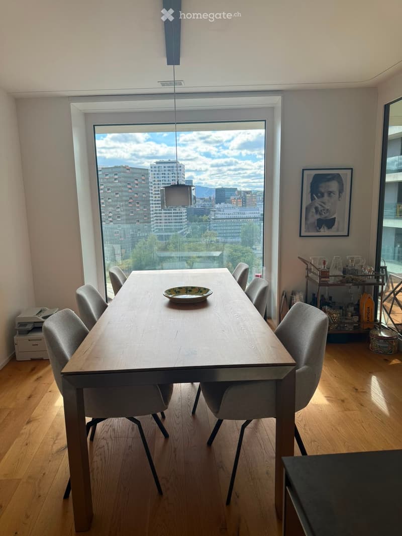 Modern dining area with a long wooden table, six chairs, a decorative plate, a small cart with bottles, and a printer near the window offering a city view.