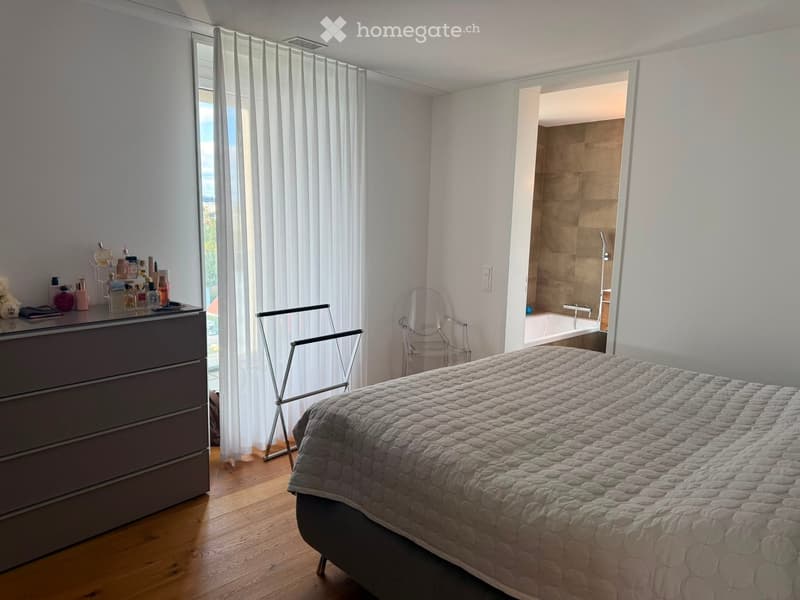 A modern bedroom with a large bed, a dresser with various items, a chair, and an open bathroom with a bathtub and shower.