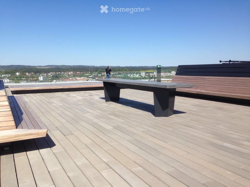 Modern rooftop terrace with wooden decking, a large rectangular table, and seating areas, offering a scenic city view.