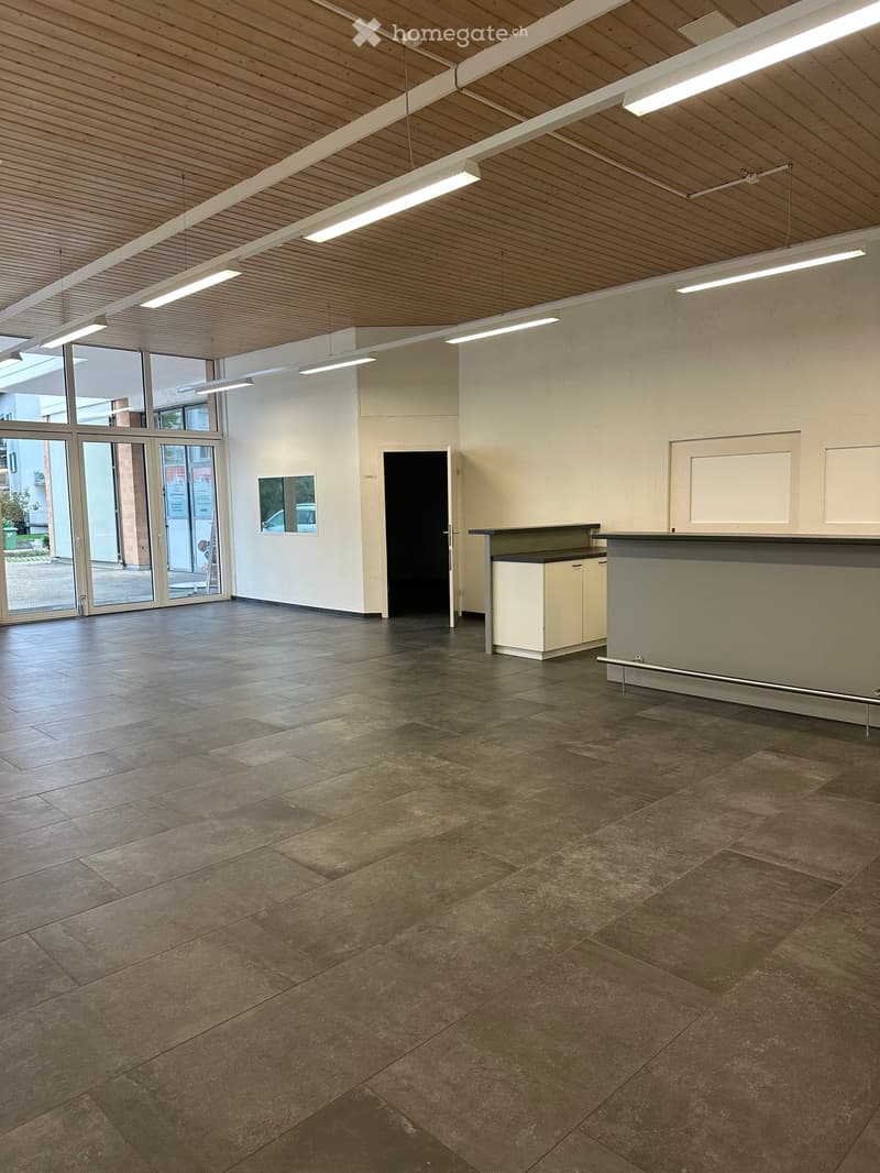 Empty room with tiled floor, white walls, and a reception desk area. Large windows and doors lead outside.