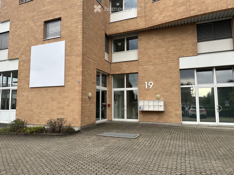 Modern multi-story building with beige brick facade, large windows, glass entrance doors, and mailboxes.