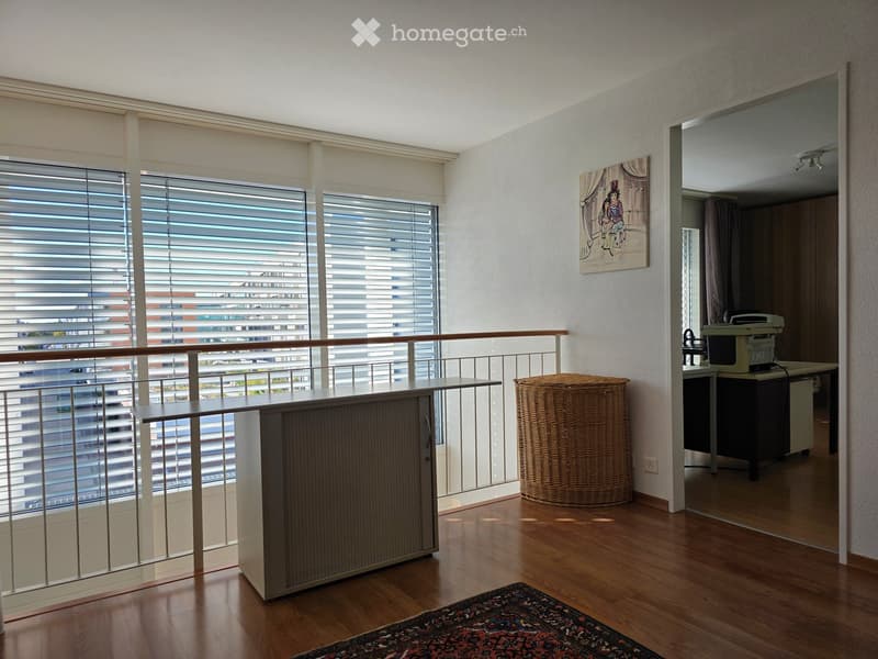 A modern, well-lit room with wooden flooring, large windows with blinds, a balcony area with a small table, a wicker basket, and a printer on a cabinet in the corner.