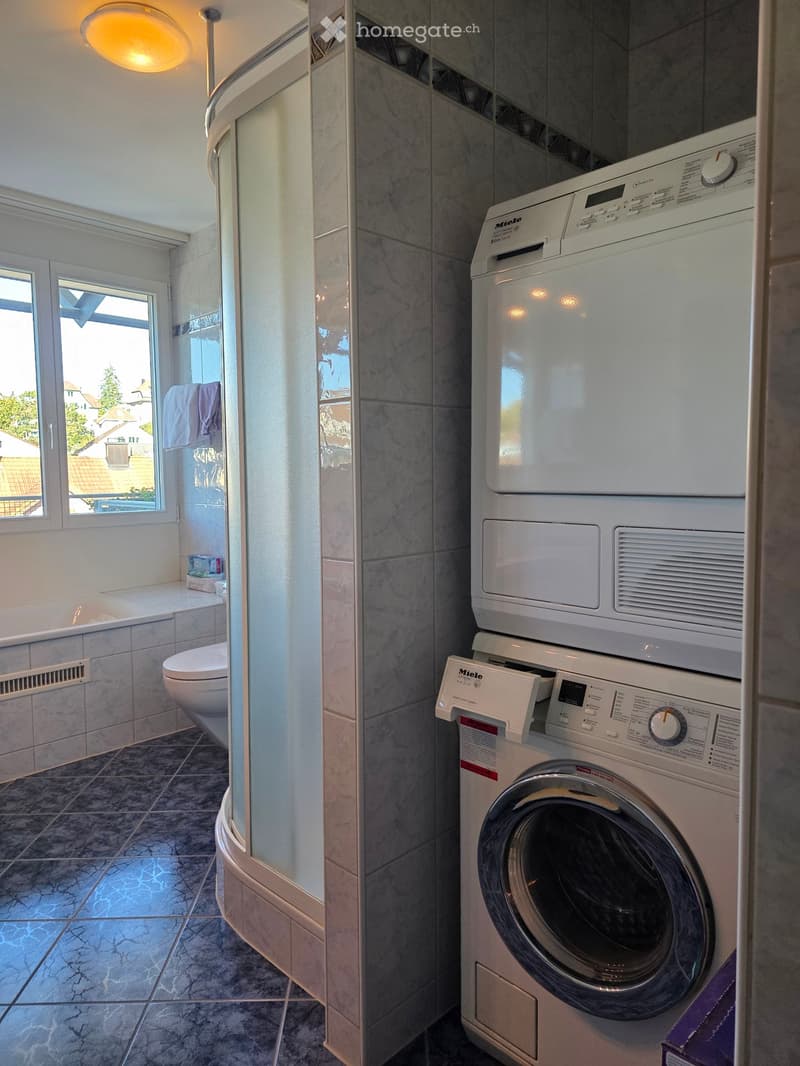 Modern bathroom with a toilet, washing machine, shower, and a towel hanging on the wall. The room features marble tiles and a window with a view of the outside.