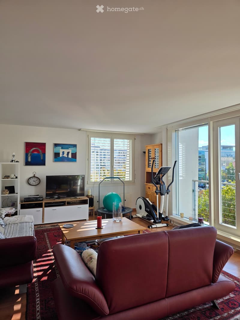 Bright living room with large windows, red leather sofa, wooden coffee table, exercise bike, TV, and artwork on the walls.