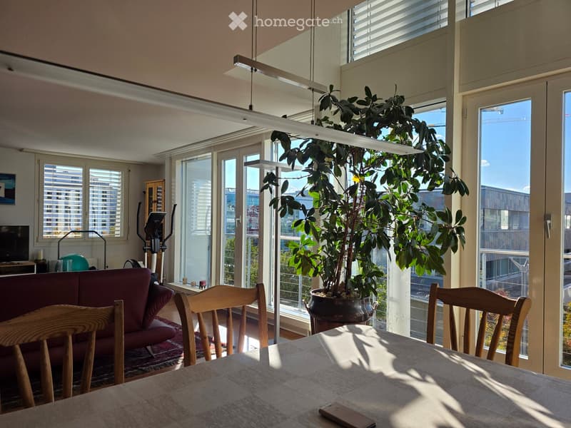 Bright living space with large windows and sliding glass doors leading to a balcony, featuring a red sofa, wooden dining table with chairs, a large potted plant, and exercise bike in the corner.