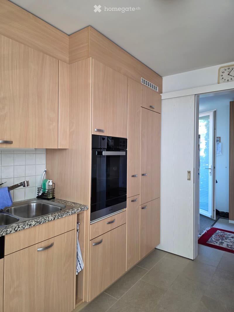 Modern kitchen with light wooden cabinets, stainless steel sink, oven, and a clock on the wall. A doorway leads to another room with a patterned rug visible.