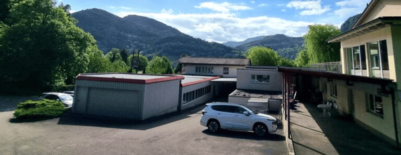 A modern building complex with a garage, parking area, and surrounding greenery, set against a scenic mountain backdrop.