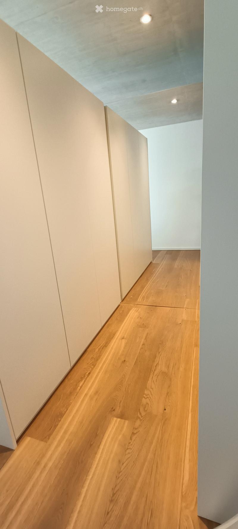 Modern hallway with light wooden flooring and white walls, featuring recessed ceiling lighting and a visible open doorway leading to another room.