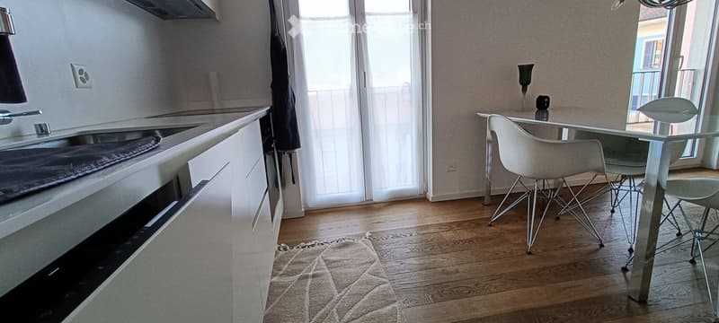 Modern kitchen with white cabinets, a kitchen island, and a dining area with a glass table and white chairs. A balcony is visible through large windows.