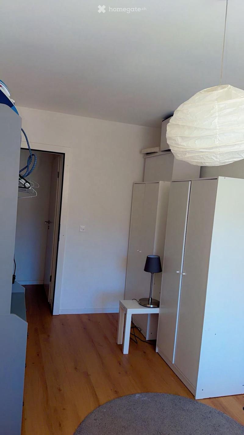 A small, unfurnished room with light wooden flooring, white walls, a small table with a lamp, and a wardrobe. The room has a grey rug on the floor and a hanging paper lantern light fixture.