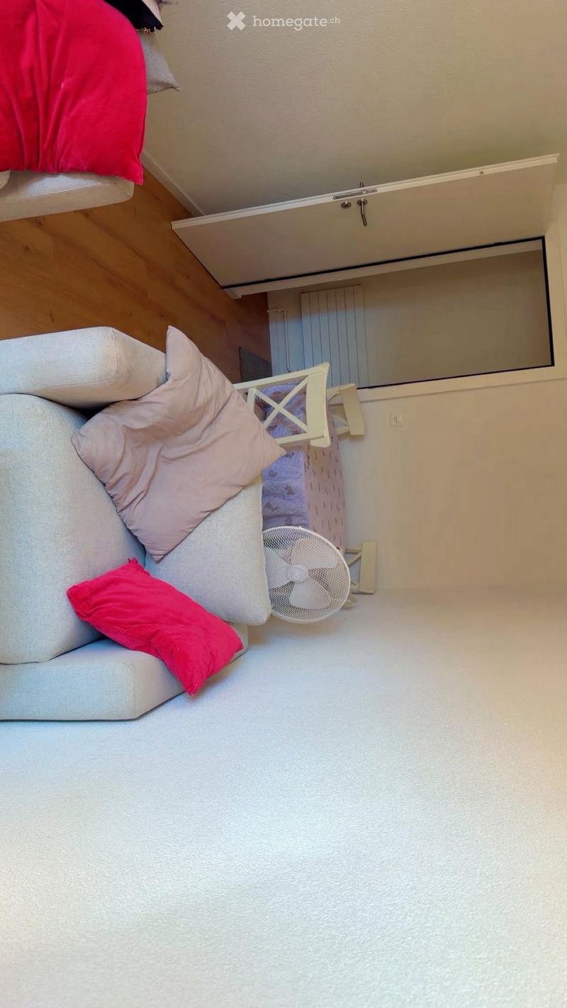 A small, cozy living space with a sofa, fan, small table, and mirror. The room features light-colored walls and a wooden floor.