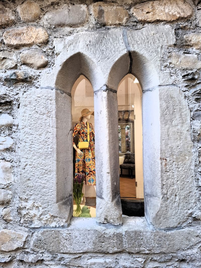 A stone wall with a unique arch window featuring a mannequin dressed in a colorful outfit and hat inside a shop, along with a vase of flowers.