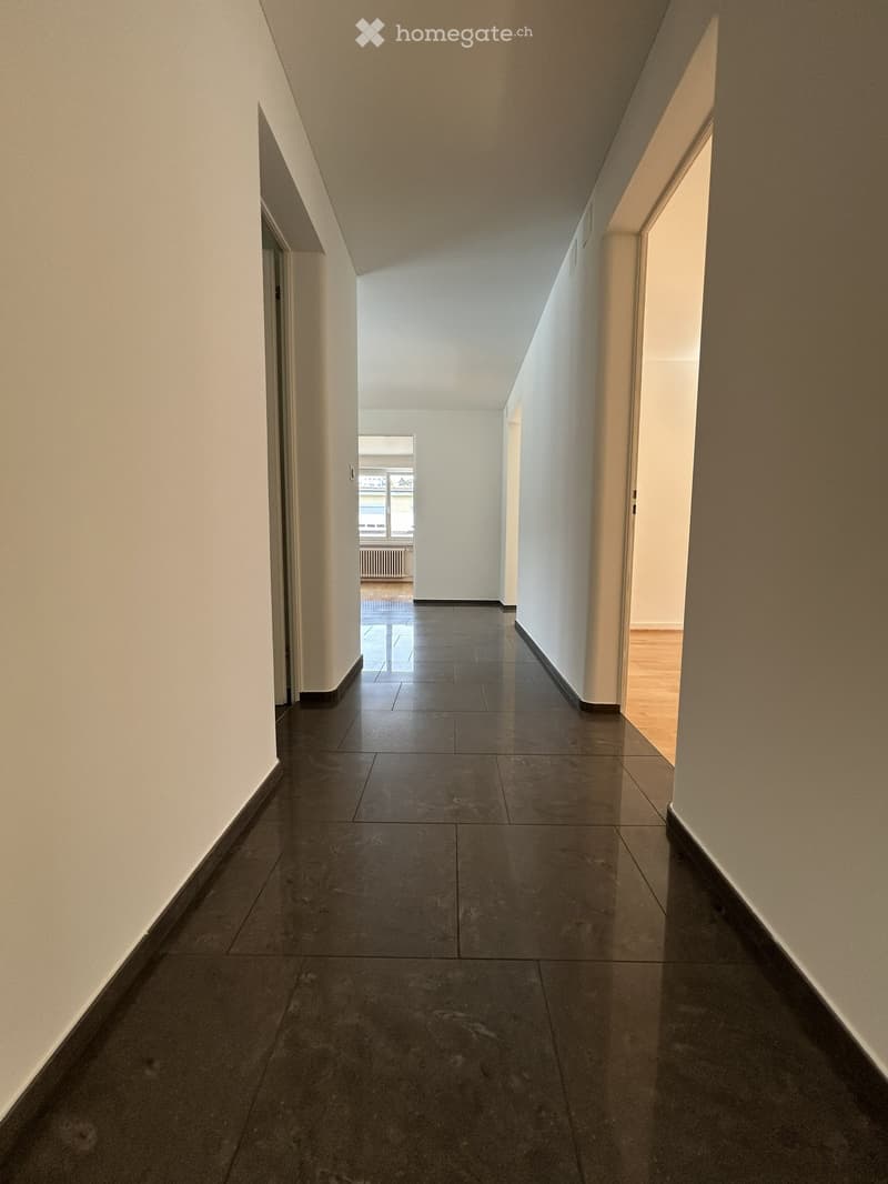 Empty hallway with dark tiled floor and white walls, leading to an open area with a radiator and window.