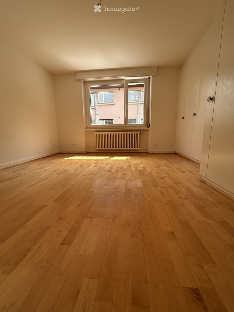 Empty room with light wooden flooring, white walls, and a large window with a radiator below it.