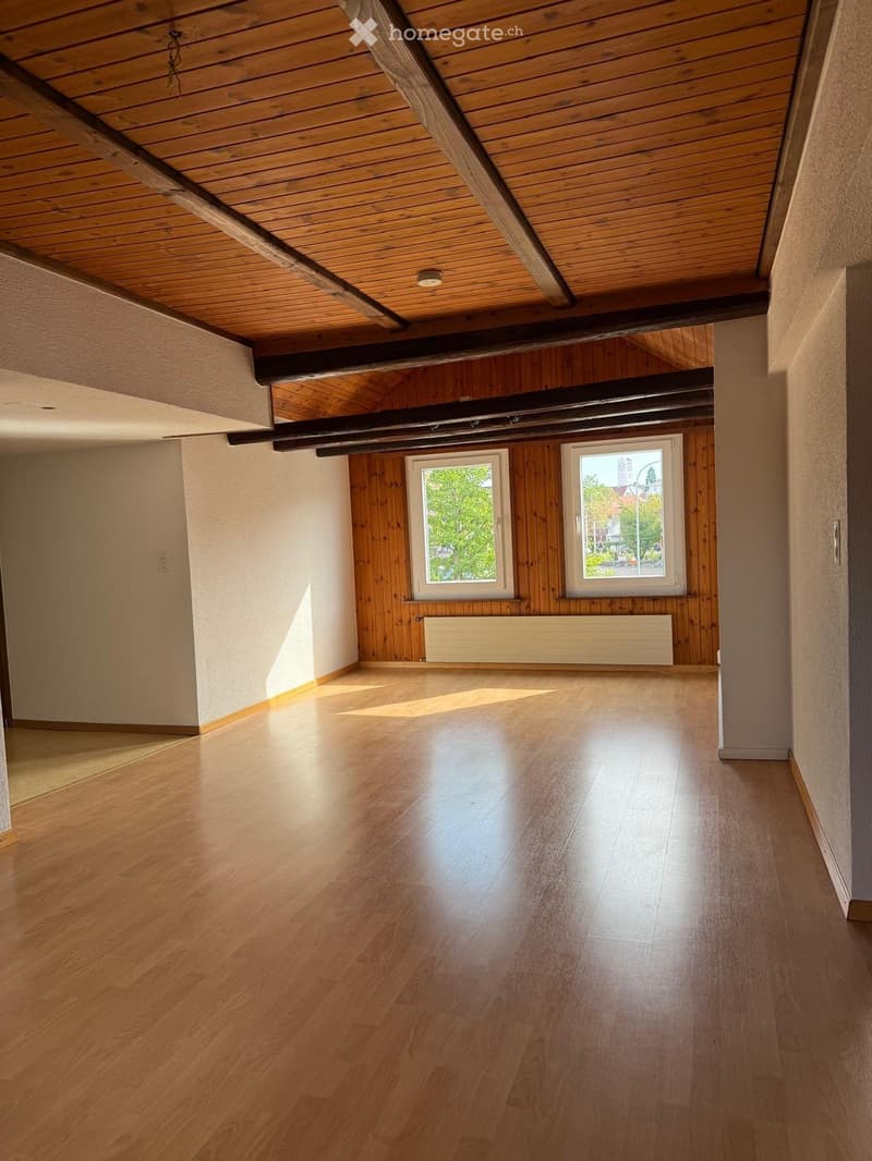 Empty room with wooden flooring, wooden walls, exposed wooden beams on the ceiling, and two windows with white frames.