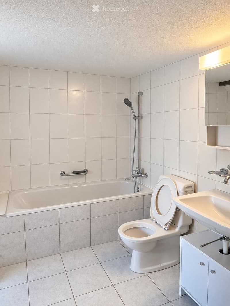 Modern bathroom with white tiled walls and floor, featuring a bathtub, toilet, sink, mirror, and showerhead.