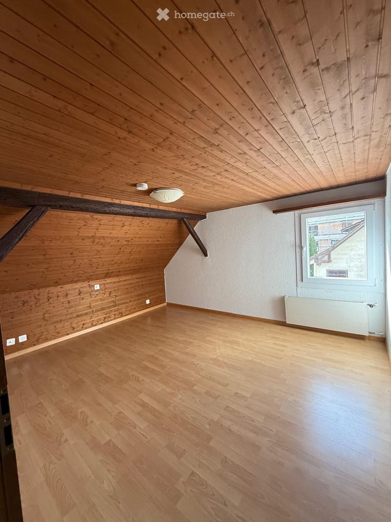 Empty room with wooden flooring and slanted wooden ceiling, featuring a window and radiator on one wall.