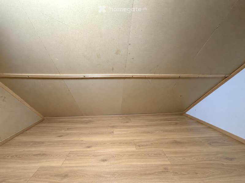 Empty room with light wooden flooring and white walls, featuring a slanted ceiling and a small window on one side.