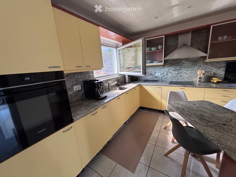 Modern kitchen with light yellow cabinets, granite countertops, and stainless steel appliances. Features an oven, coffee maker, sink, and a small dining area with a table and chairs.