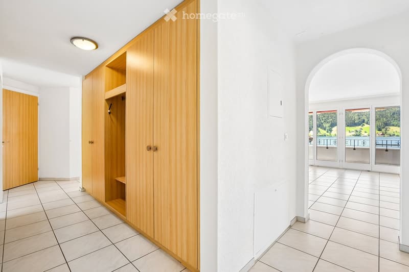 Bright hallway with tiled floor, white walls, wooden built-in closet, and an arched opening leading to a balcony with scenic views.