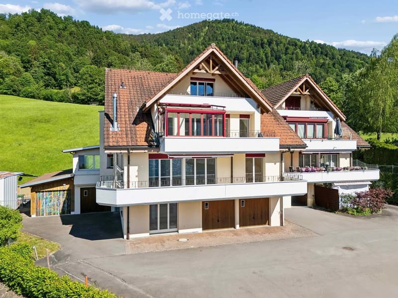 Two-story house with a tiled roof, large windows, balconies, and a garage. Surrounded by greenery and a driveway.