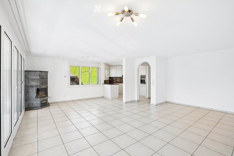 Bright, open living space with tiled flooring, a fireplace, a kitchen area with a white island, and large windows offering a view of greenery.