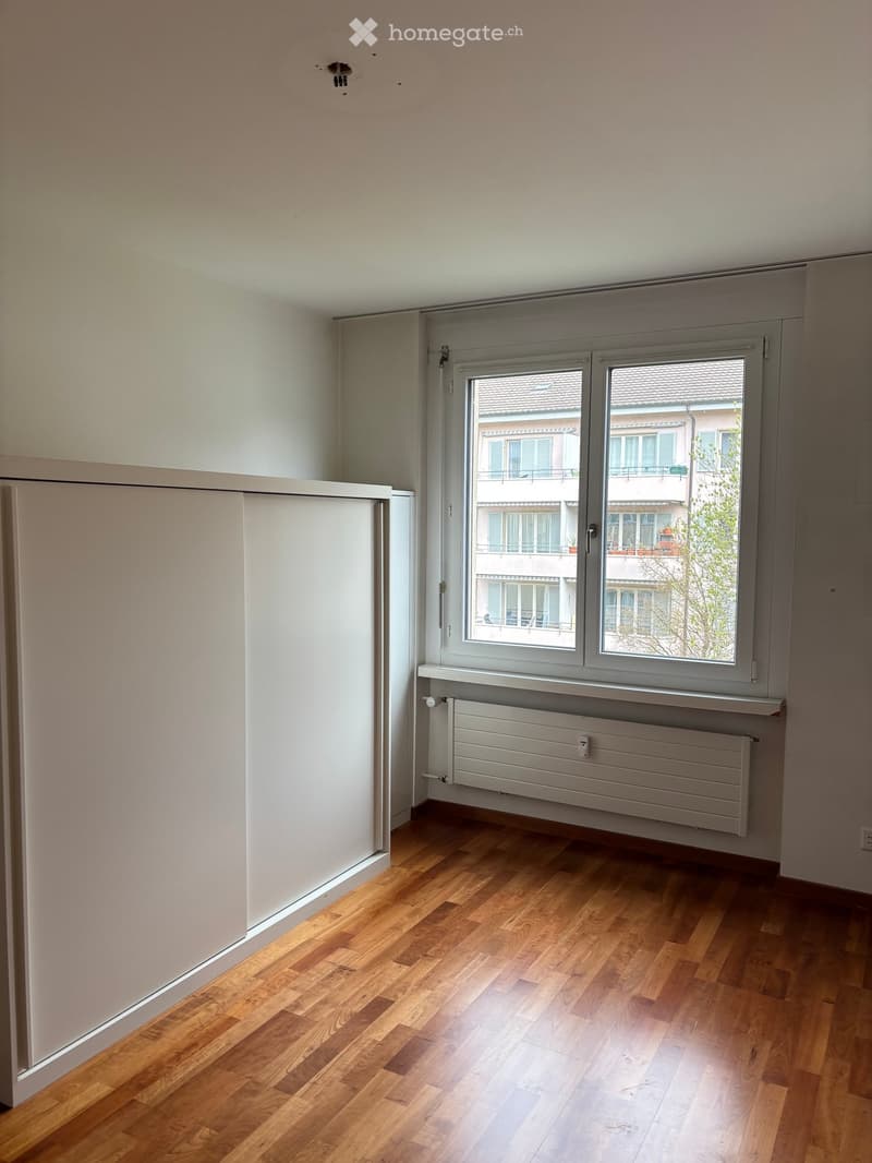 Empty room with wooden flooring, white walls, and a large window with a radiator below it. The window offers a view of neighboring buildings.
