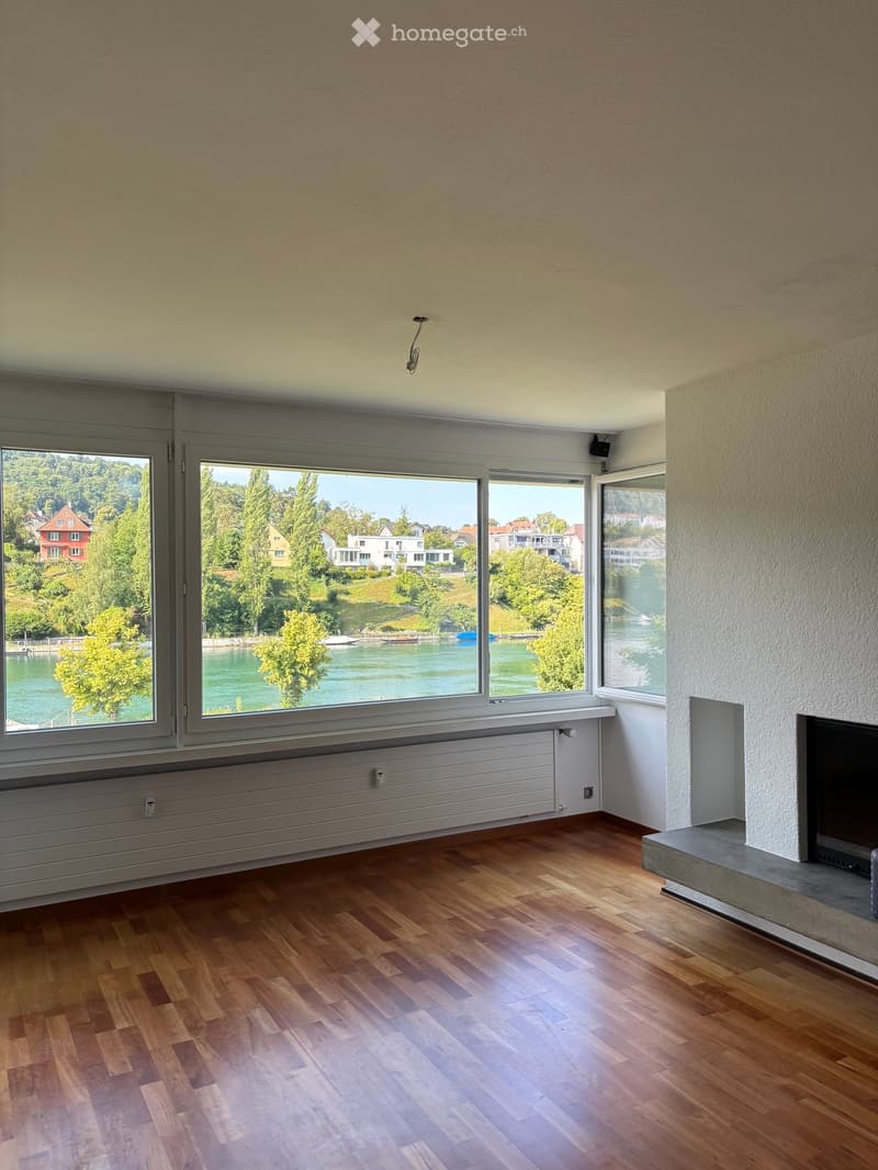 Empty room with wooden flooring, large windows offering a scenic view of a lake and surrounding houses, and a radiator below the windows.