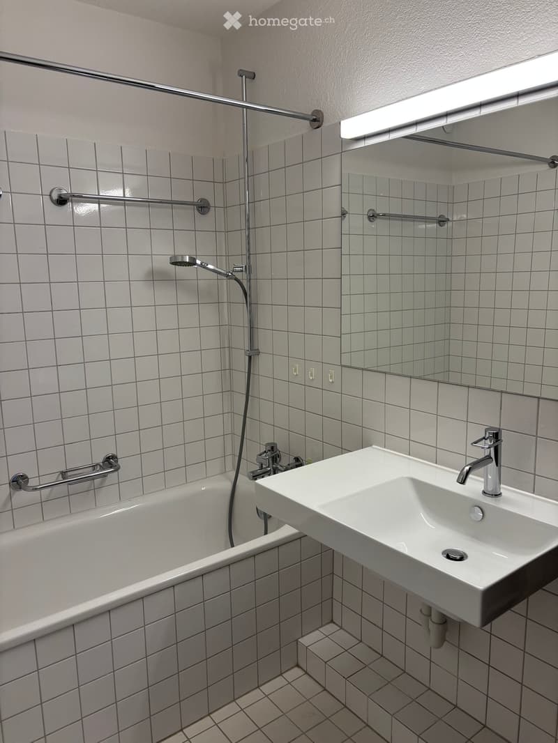 Modern bathroom with white tiled walls and floor, featuring a bathtub, a wall-mounted sink with a modern faucet, a showerhead, and a large mirror above the sink.