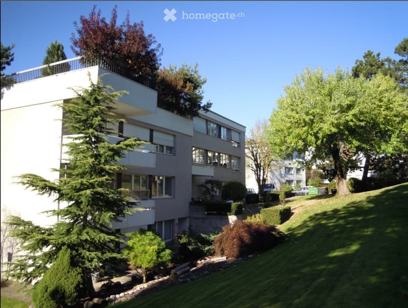 Modern multi-story building with balconies, surrounded by trees and greenery on a hillside.
