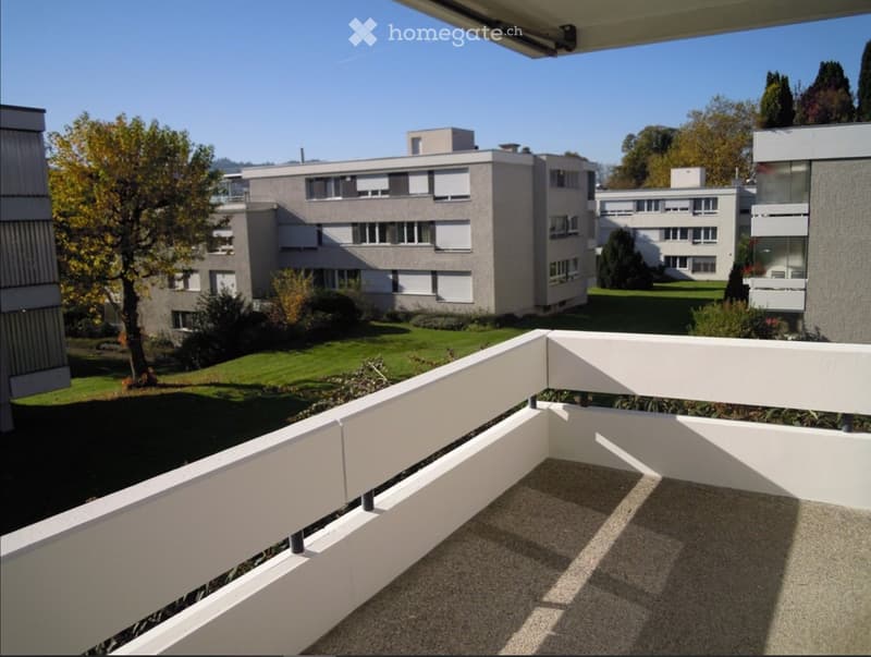 Balcony with white railing overlooking a landscaped area with modern buildings and trees.