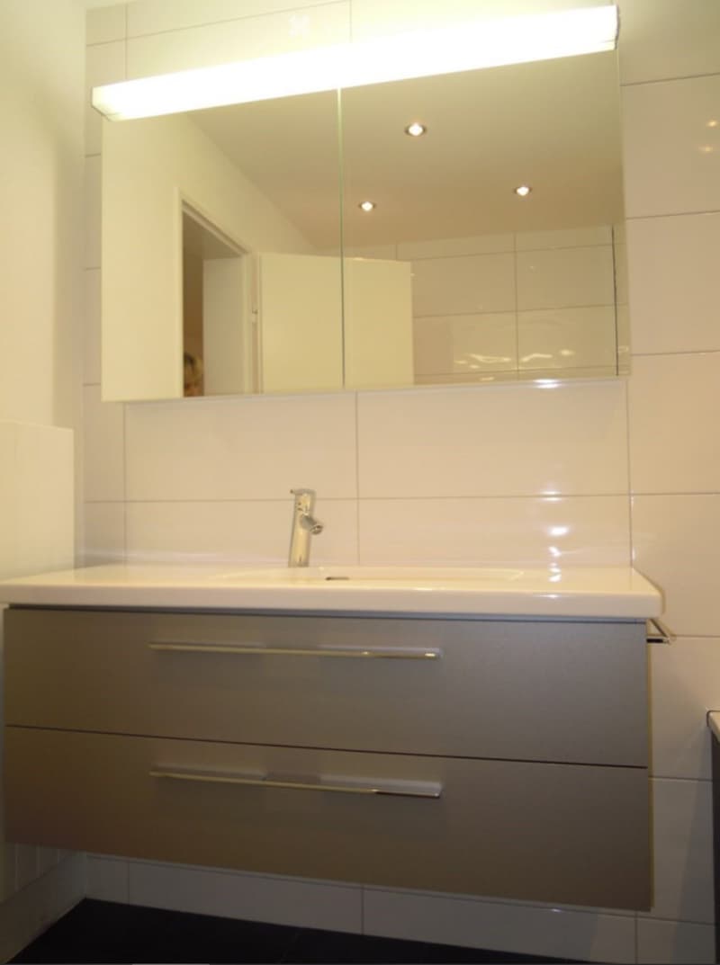 Modern bathroom with a white sink, chrome faucet, large mirror with integrated lighting, and a grey cabinet with silver handles.