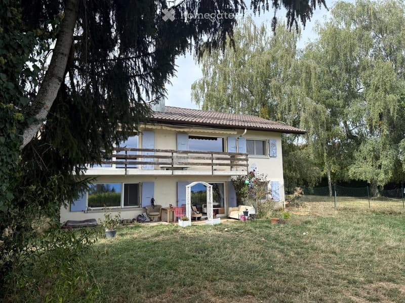Two-story house with a tiled roof, light-colored walls, and multiple windows. Features a wooden balcony on the upper level and a small outdoor seating area with chairs and a table in the front yard.