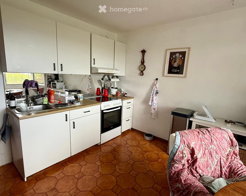 The kitchen features white cabinets, a sink, an oven, and a microwave. There is a small table with a pink blanket on it, a picture frame on the wall, and a clock. The floor is tiled in a hexagonal pattern.